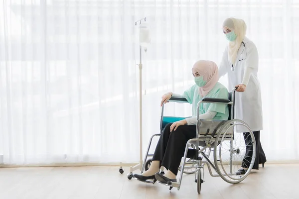 ヒジャーブのドレスを着たイスラム教徒の女性医師は親切に病院の車椅子に座っている女性患者を助けます 彼らはウイルスや病気から彼らを守るために外科衛生保護マスクを顔につけています — ストック写真