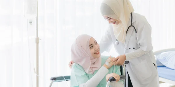 Attractive young beautiful female Muslim doctor covered face with hijab dress talking and kindly take care for woman patient who sitting on a wheelchair in the hospital. Idea for health care business.