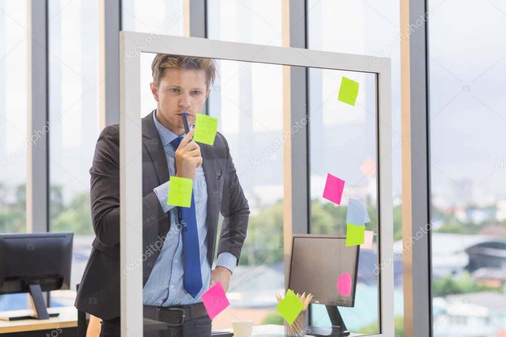 Handsome Caucasian businessman looking at sticky note on glass board and find some good idea from it. This is an idea from coworker brainstorming.