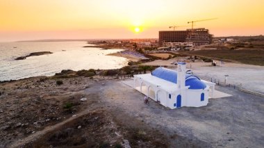 Kıbrıs Rum Kesimi 'nin Protaras, Famagusta, Agia Triada plajındaki kıyı şeridi günbatımı ve tarihi beyaz şapel manzarası. Gündoğumunda Ammochostos ilçesinde Ayia Trias Körfezi Kilisesi, Kum ve liman turistik bölge kuşbakışı.