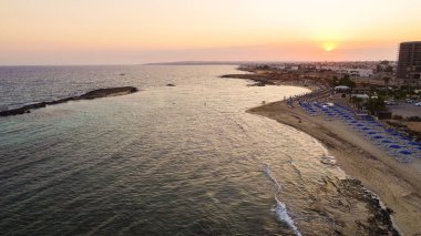 Kıyı şeridi günbatımı ve tarihi plaj manzarası yukarıdan Agia Thekla, Ayia Napa, Famagusta, Kıbrıs. Kuş bakışı gökyüzü manzarası turistlerin ilgi odağı altın kumlu körfez, adacık, Ammochostos 'taki güneşli yataklar..