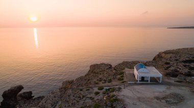 Aerial view of coastline sunset and landmark white washed chapel Agioi Anargyroi, at Cavo Greco Protaras, Famagusta, Cyprus from above. Bird's eye view of tourist attraction cliff rock Ayioi Anargiroi church, caves, beach at sunrise in Ammochostos di clipart