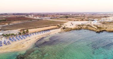 Kıyı şeridi günbatımı ve tarihi plaj manzarası yukarıdan Agia Thekla, Ayia Napa, Famagusta, Kıbrıs. Kuş bakışı gökyüzü manzarası turistlerin ilgi odağı altın kumlu körfez, adacık, Ammochostos 'taki güneşli yataklar..
