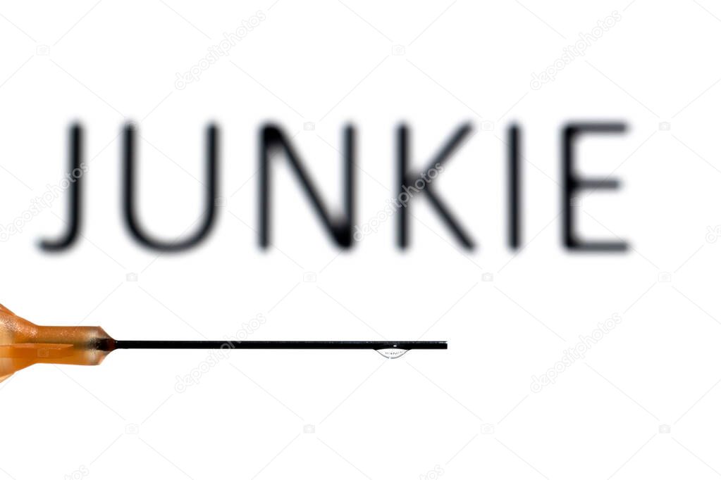 A medical needle with a droplet suspended refracting the word junkie, which is also out of focus in the background