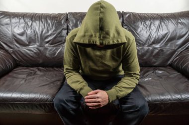 Man in dark green hoodie with unrecognizable face sitting on broun leather sofa