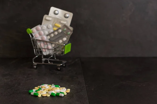 Supermarket cart with various pills and medicines on a black background. Around the cart are scattered tablets and capsules of different colors. Coronavirus protection. A lot of multi-colored tablets