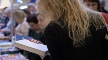 Vilnius, Litvanya - Şubat 2020 - Kitap Fuarı 'nda diğer insanlarla çevrili bir kitap tanımı okuyan genç kadın