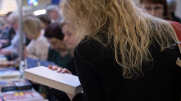 Vilnius, Litouwen - februari 2020 - Jonge vrouw die een boekbeschrijving leest in Book Fair omringd door andere mensen — Stockvideo