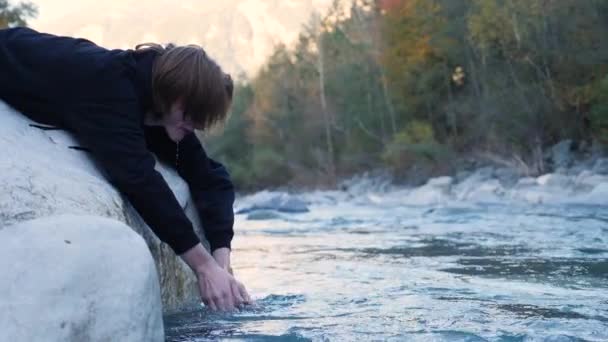 Joven hombre de pelo largo se lava la cara con agua fría del río de manantial de montaña acostado en la piedra por la mañana — Vídeos de Stock