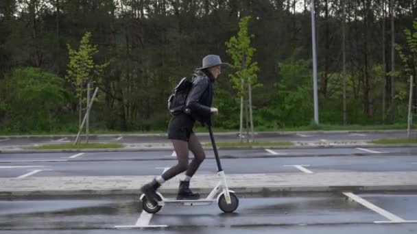 Jonge nomade vrouw met rugzak rijdt elektrische scooter. Vrouw draagt hoed en laarzen, neergeschoten op parkeerplaats in de buurt van bos. — Stockvideo