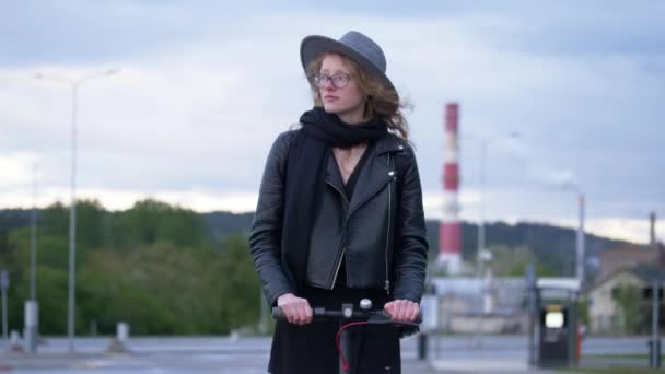 Kaukasisch aussehende Frau fährt Elektroroller und bewundert die Umwelt. Trägt Hut und Brille. Parkplatz im Hintergrund. — Stockvideo