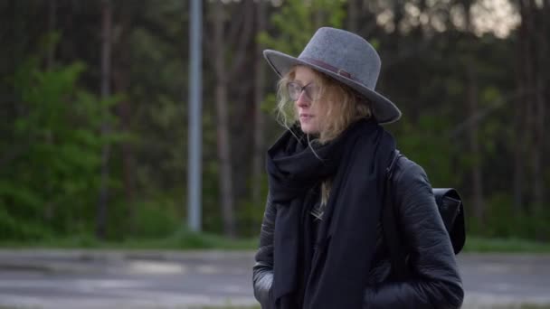 若い濃縮された女性はまっすぐ歩く。ブラックカラーにこだわり、帽子と眼鏡をかけています。背景の森. — ストック動画