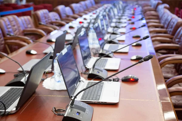 In de grote hal, een lange tafel van hout voor onderhandelingen, open computers en microfoons aan tafel zijn bruine stoelen. bedrijfsconcept. — Stockfoto