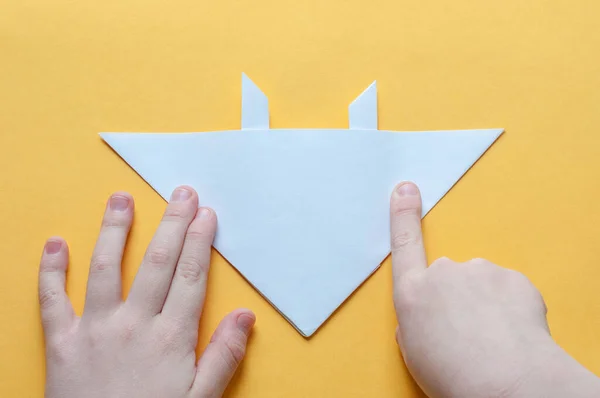 15. adım. Sarı arka plandaki bir çocuğun elleri beyaz bir kağıdın karesini üçgenlere böler. Origami boğa kavramı.