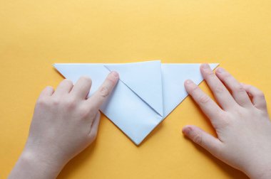 Altıncı adım. Sarı arka plandaki bir çocuğun elleri beyaz bir kağıdın karesini üçgenlere böler. Origami boğa kavramı.