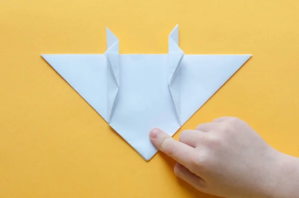 Step 14. Hands of a child on a yellow background divide a square of white paper into triangles. Origami bull concept.