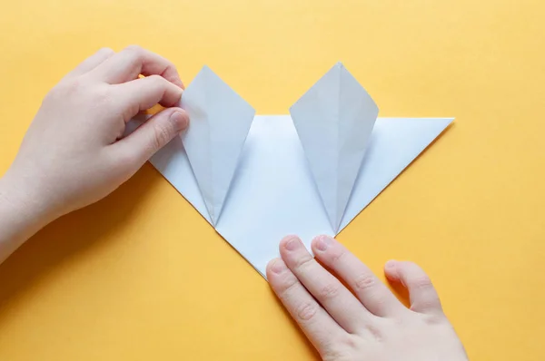 11. adım. Sarı arka plandaki bir çocuğun elleri beyaz bir kağıdın karesini üçgenlere böler. Origami boğa kavramı.