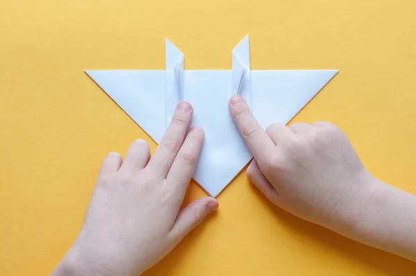 13. adım. Sarı arka plandaki bir çocuğun elleri beyaz bir kağıdın karesini üçgenlere böler. Origami boğa kavramı.