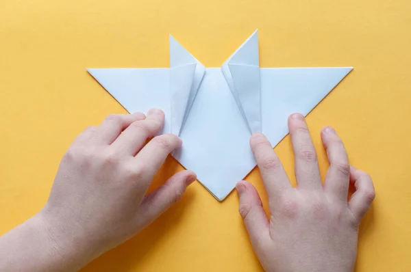 12. adım. Sarı arka plandaki bir çocuğun elleri beyaz bir kağıdın karesini üçgenlere böler. Origami boğa kavramı.
