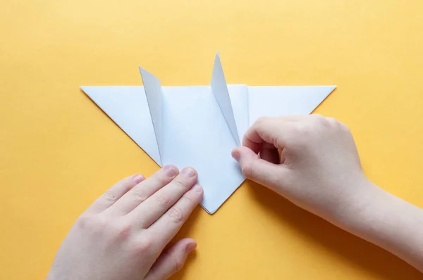 9. adım. Sarı arka plandaki bir çocuğun elleri beyaz bir kağıdın karesini üçgenlere böler. Origami boğa kavramı.