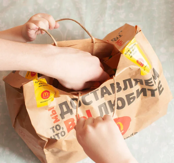 Russland, Moskau, April 2020: Auf dem Tisch liegt eine Packpapiertüte für McDonalds, Kinderhände öffnen die Tüte für selbstgemachtes Essen. Lieferkonzept Illustrative Version der Ware. — Stockfoto