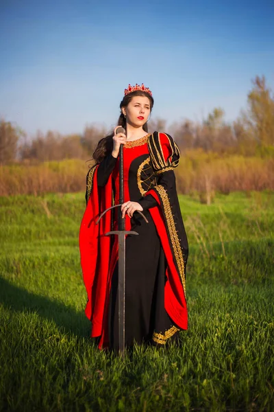 一个身穿中世纪黑色和红色连衣裙 头戴皇冠的年轻漂亮女子站在田野里 手里拿着一把剑 中世纪女王的概念艺术手里拿着武器 寂寞的公主在田野里散步 — 图库照片