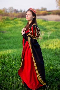 Kafasında taç olan siyah ve kırmızı ortaçağ elbisesi içindeki genç ve güzel bir kadın bir tarlada duruyor. Prenses akşam yeşil çimlerde yürür.