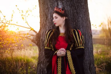 Çekici, siyah ve kırmızı ortaçağ elbiseli, başında taç olan bir kız büyük bir ağacın arkasında duruyor. 14-15 yüzyıl tarzında giyinmiş genç bir kraliçe..