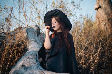 Soluk tenli, siyah dudaklı, siyah şapkalı, elbiseli ve pelerinli genç bir cadı. Sonbaharda, devrilmiş bir ağacın arka planında, sarı, kurumuş çimenler ve mavi gökyüzü. Cadılar bayramı, sihir, fantezi imajı.