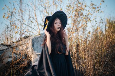 Soluk tenli, siyah dudaklı, siyah şapkalı, elbiseli ve pelerinli genç bir cadı. Sonbaharda, devrilmiş bir ağacın arka planında, sarı, kurumuş çimenler ve mavi gökyüzü. Cadılar bayramı, sihir, fantezi imajı.