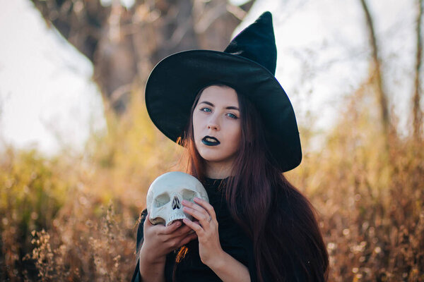 A young witch with pale skin and black lips in a black hat, a dress and a raincoat. Holds the skull of a dead person. Against the background of a tree of gallows. halloween, magic, fantasy, necromancy.