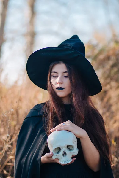 一个年轻的巫婆 苍白的皮肤 黑色的嘴唇 戴着黑色的帽子 穿着连衣裙和雨衣 拿着一个死人的骷髅在一棵绞刑架树的后面神圣的 魔法的 幻想的 死灵的 — 图库照片