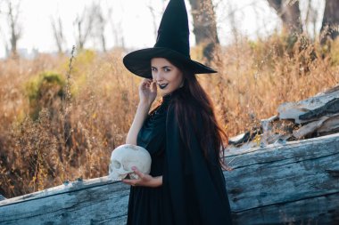 Soluk tenli, siyah dudaklı, siyah şapkalı, elbisesi ve yağmurluğu olan genç bir cadı. Ölü bir adamın kafatasını tutuyor. Sonbahar, devrilmiş ağaç ve uzun, kuru otlar. Cadılar Bayramı, sihir, fantezi.