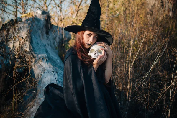 A young witch with pale skin and black lips in a black hat, a dress and a raincoat. Holds the skull of a dead man. Autumn, fallen tree and tall, dry grass. Halloween, magic, fantasy.