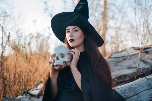 A young witch with pale skin and black lips in a black hat, a dress and a raincoat. Holds the skull of a dead man. Autumn, fallen tree and tall, dry grass. Halloween, magic, fantasy.
