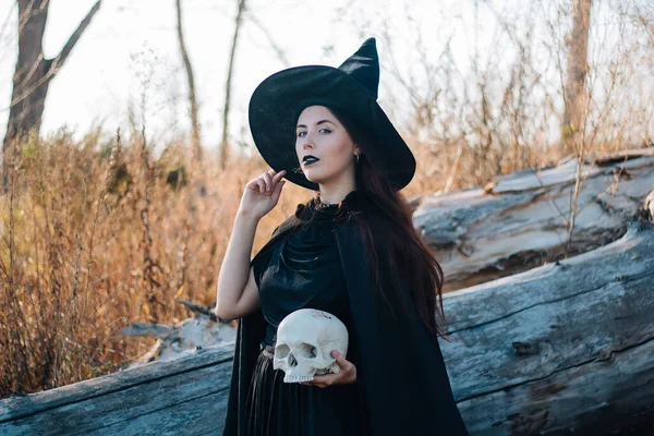 黒い帽子 ドレス レインコートに薄い肌と黒い唇を持つ若い魔女 死んだ男の頭蓋骨を持ってる 倒木や背の高い 乾燥した草 ハロウィン魔法ファンタジー — ストック写真