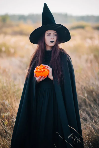 一个年轻的巫婆 苍白的皮肤 黑色的嘴唇 戴着黑色的帽子 穿着衣服和斗篷 手里拿着南瓜 穿着女巫服装的女孩在万圣节 秋天在田野的背景下 — 图库照片