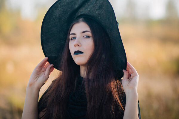 Portrait of a beautiful girl with pale skin and black lips in a big black hat. Woman in the image of a witch for Halloween.