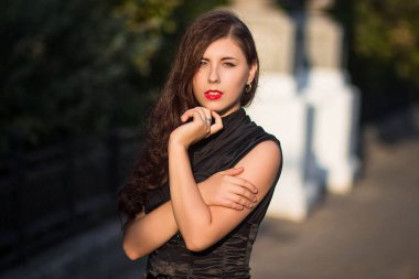 Korseli siyah elbiseli genç, güzel bir kadın, şehir caddesinde arka planda bir park ve fenerli taş sütunlarla yürüyor..