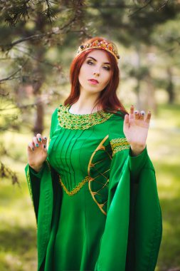 Yeşil, genç, kızıl saçlı, uzun kollu, yeşil bir ortaçağ elbisesi içinde altın bir tacı olan, bir peri ormanında çimlerin üzerinde duran güzel bir kız. Muhteşem bir fotodurgunluk. Temiz ciltli bir model..