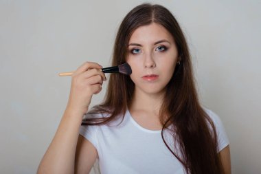 Beyaz tişörtlü genç, güzel bir kız yüzüne makyaj yapıyor kozmetik fırçasıyla. Beyaz arkaplanda stüdyo fotoğrafı.