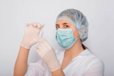 Kadın bir doktorun elinde bir aşı ampulünün yakın çekimi var. Beyaz önlüklü, tıbbi şapkalı, maskeli ve eldivenli laboratuvar teknisyeni elinde ilaç tutuyor. Coronavirus aşısı arama konsepti.