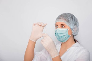 Kadın bir doktorun elinde bir aşı ampulünün yakın çekimi var. Beyaz önlüklü, tıbbi şapkalı, maskeli ve eldivenli laboratuvar teknisyeni elinde ilaç tutuyor. Coronavirus aşısı arama konsepti.