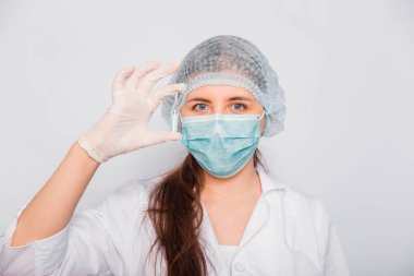 Kadın bir doktorun elinde bir aşı ampulünün yakın çekimi var. Beyaz önlüklü, tıbbi şapkalı, maskeli ve eldivenli laboratuvar teknisyeni elinde ilaç tutuyor. Coronavirus aşısı arama konsepti.