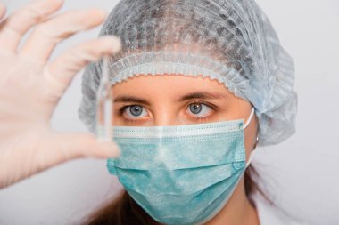 Kadın bir doktorun elinde bir aşı ampulünün yakın çekimi var. Beyaz önlüklü, tıbbi şapkalı, maskeli ve eldivenli laboratuvar teknisyeni elinde ilaç tutuyor. Coronavirus aşısı arama konsepti.
