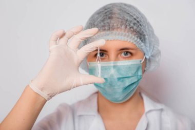 Beyaz önlüklü, tıbbi şapkalı, maskeli ve eldivenli bir kadın doktor aşı ile bir ampul tutuyor. Coronavirus aşısı arama konsepti.
