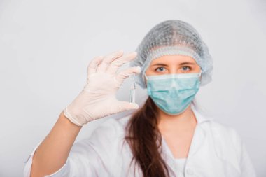 Beyaz önlüklü, tıbbi şapkalı, maskeli ve eldivenli bir kadın doktor aşı ile bir ampul tutuyor. Coronavirus aşısı arama konsepti.
