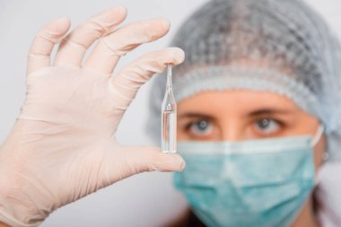 Beyaz önlüklü, tıbbi şapkalı, maskeli ve eldivenli bir kadın doktor aşı ile bir ampul tutuyor. Coronavirus aşısı arama konsepti.