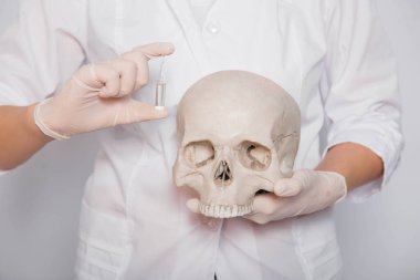 Beyaz önlüklü ve eldivenli bir doktorun ellerinde aşı ile insan kafatası ve ampul bulundu. Beyaz arka planda duruyor. Coronavirus aşısı arama konsepti.