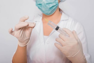 Beyaz önlüklü, tıbbi şapkalı, maskeli ve eldivenli kadın doktor şırınga iğnesi ve tıbbi ampulle çalışıyor. Steril giysiler içindeki bir bilim adamı beyaz arka planda duran bir aşı hazırlıyor. Coronavirus aşısı arama konsepti.